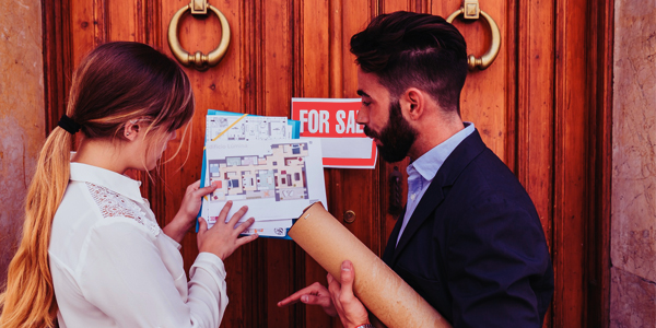 A picture of a person holding a “For Sale” sign with a real estate agent standing next to them. The background could be a house or a neighborhood.